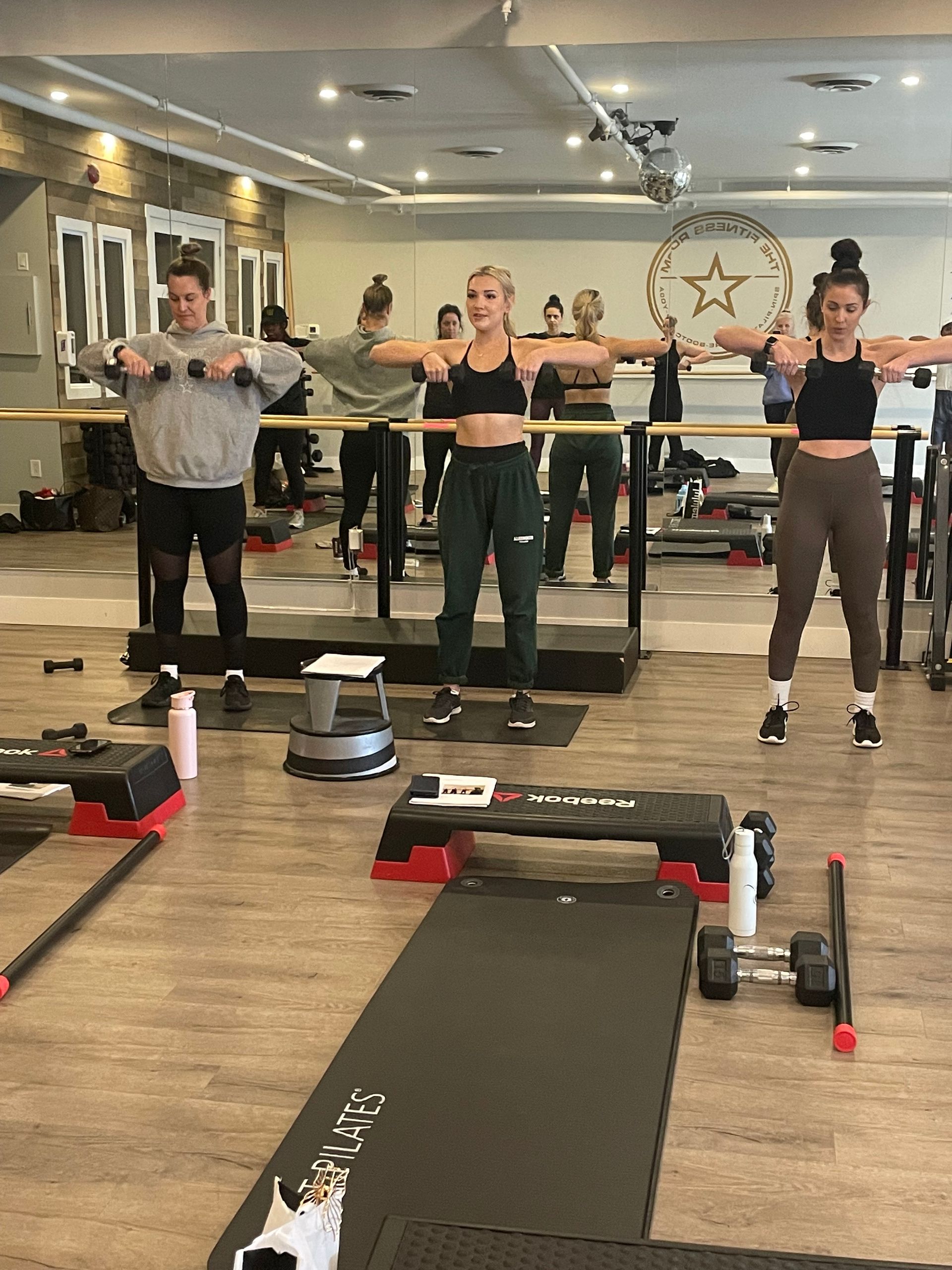 People exercising in a mirrored fitness studio, standing on mats with arms extended.