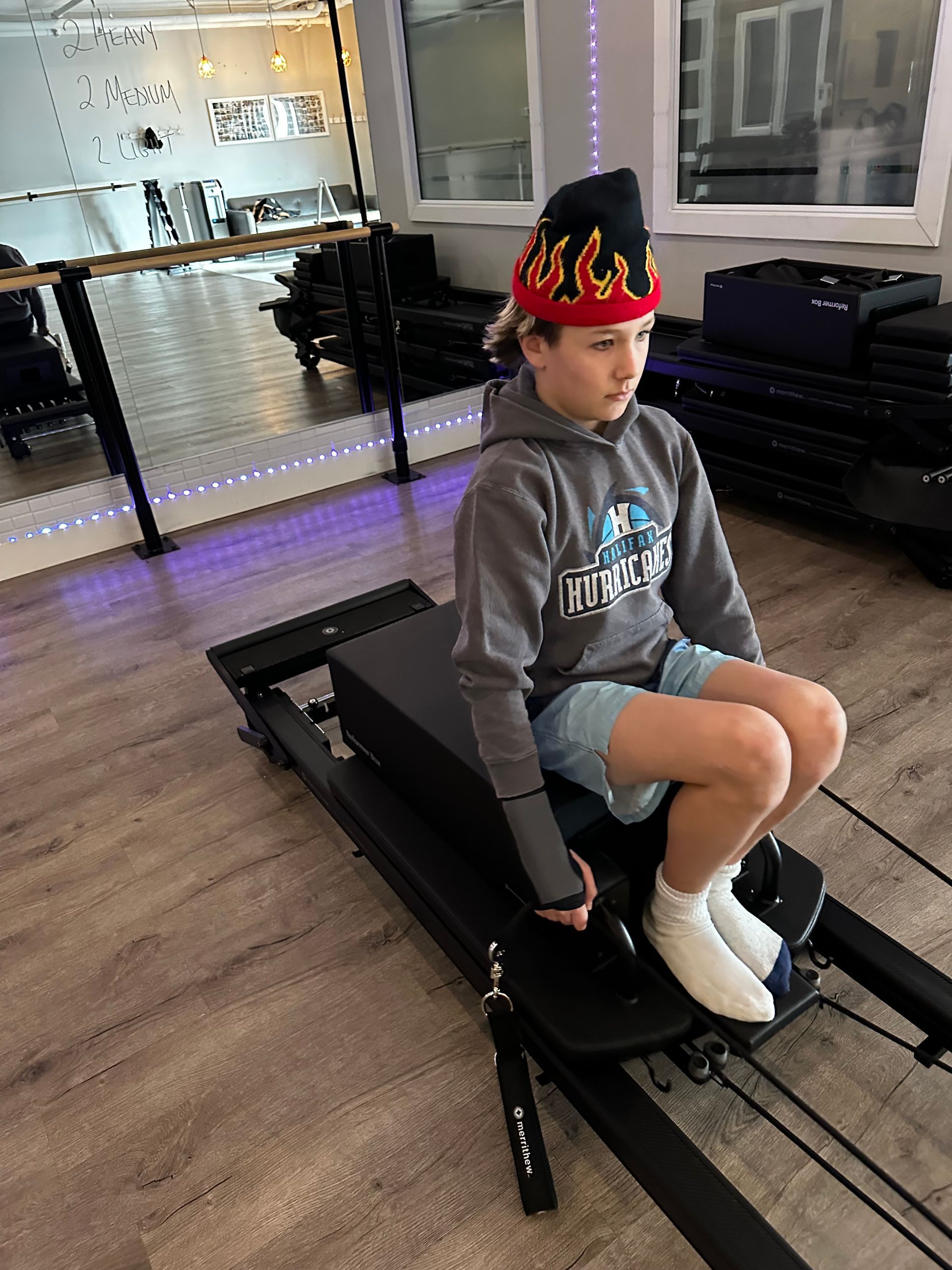 Child in a gray hoodie and flame hat sitting on a Pilates reformer in a studio.