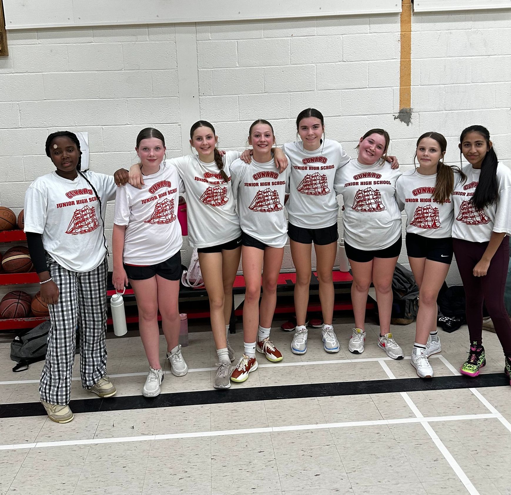 Eight teammates in white shirts and shorts pose together in a gym, smiling after a sports event.