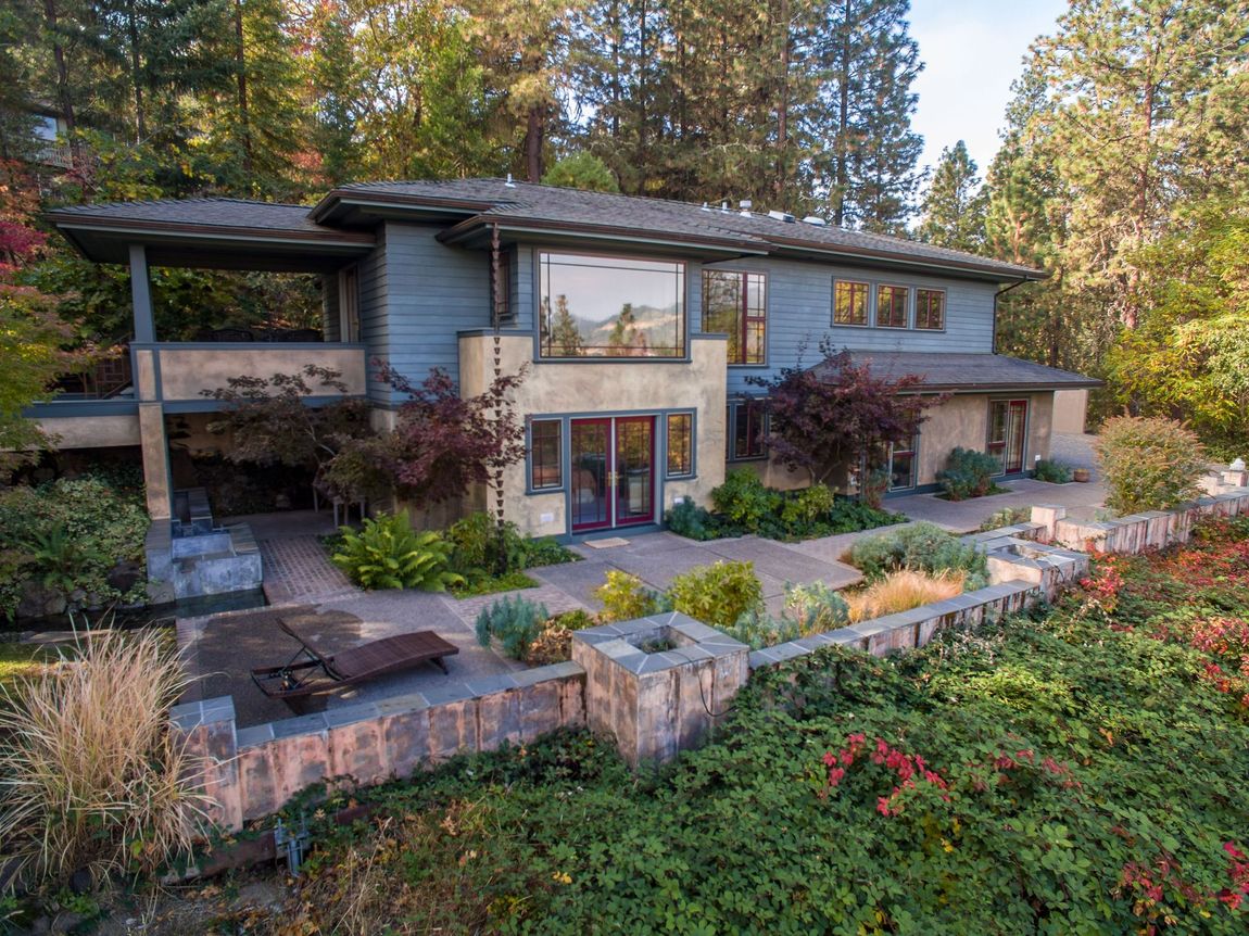 Modern two-story house with stone patio, large windows, and landscaped garden in a wooded setting