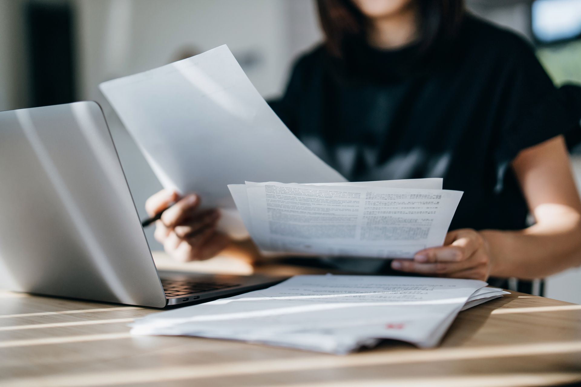 A woman is sitting at a table holding a piece of paper in front of a laptop computer.