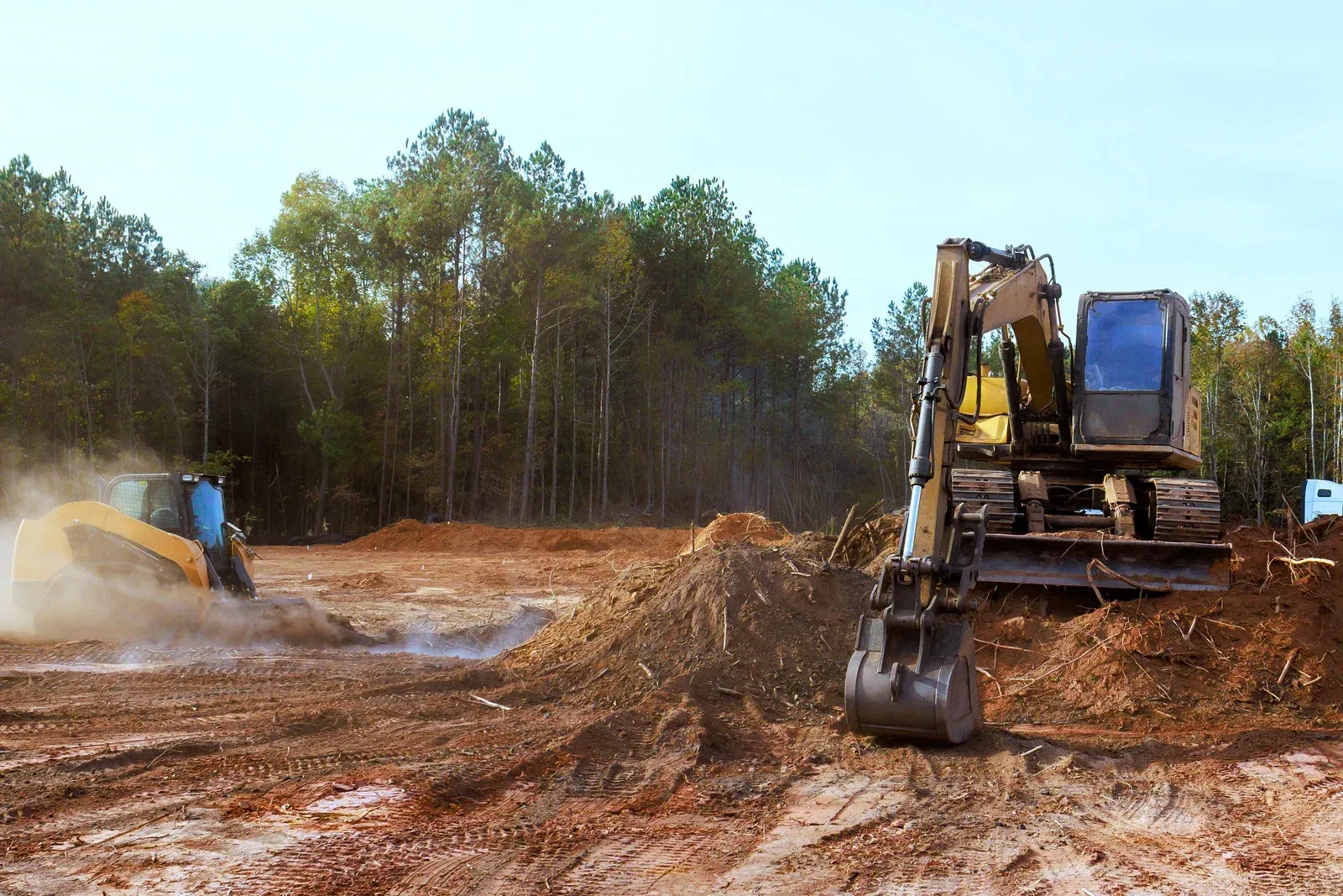 Two yellow excavators clearing muddy land near a wooded area under a cloudy sky