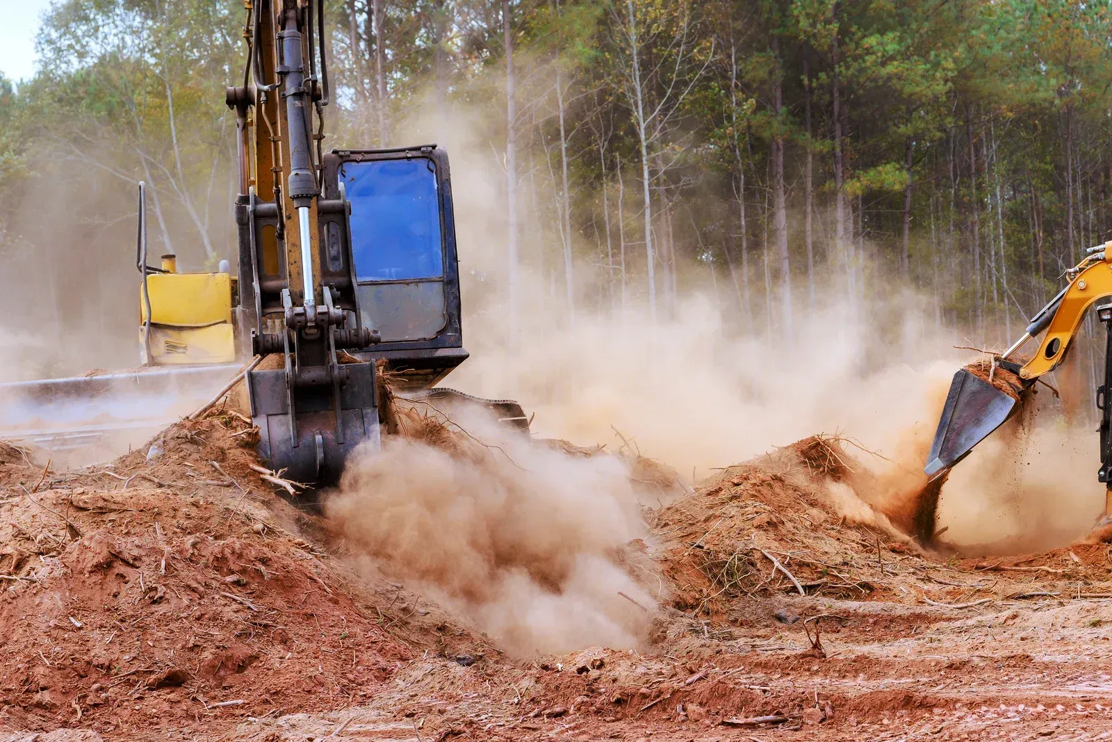 Excavator digging red dirt on a dusty construction site with trees in the background