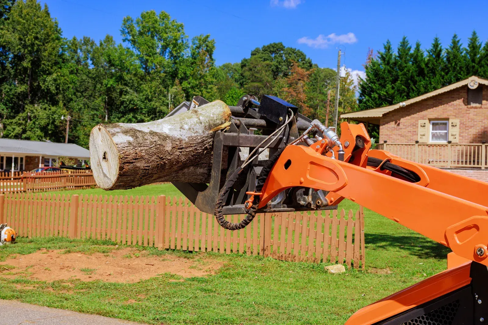Orange excavator lifting a large tree trunk in a grassy yard near a house and fence