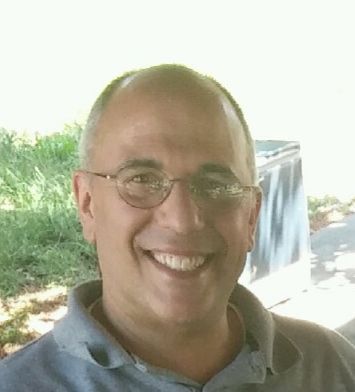 A man wearing glasses and a grey shirt is smiling for the camera.