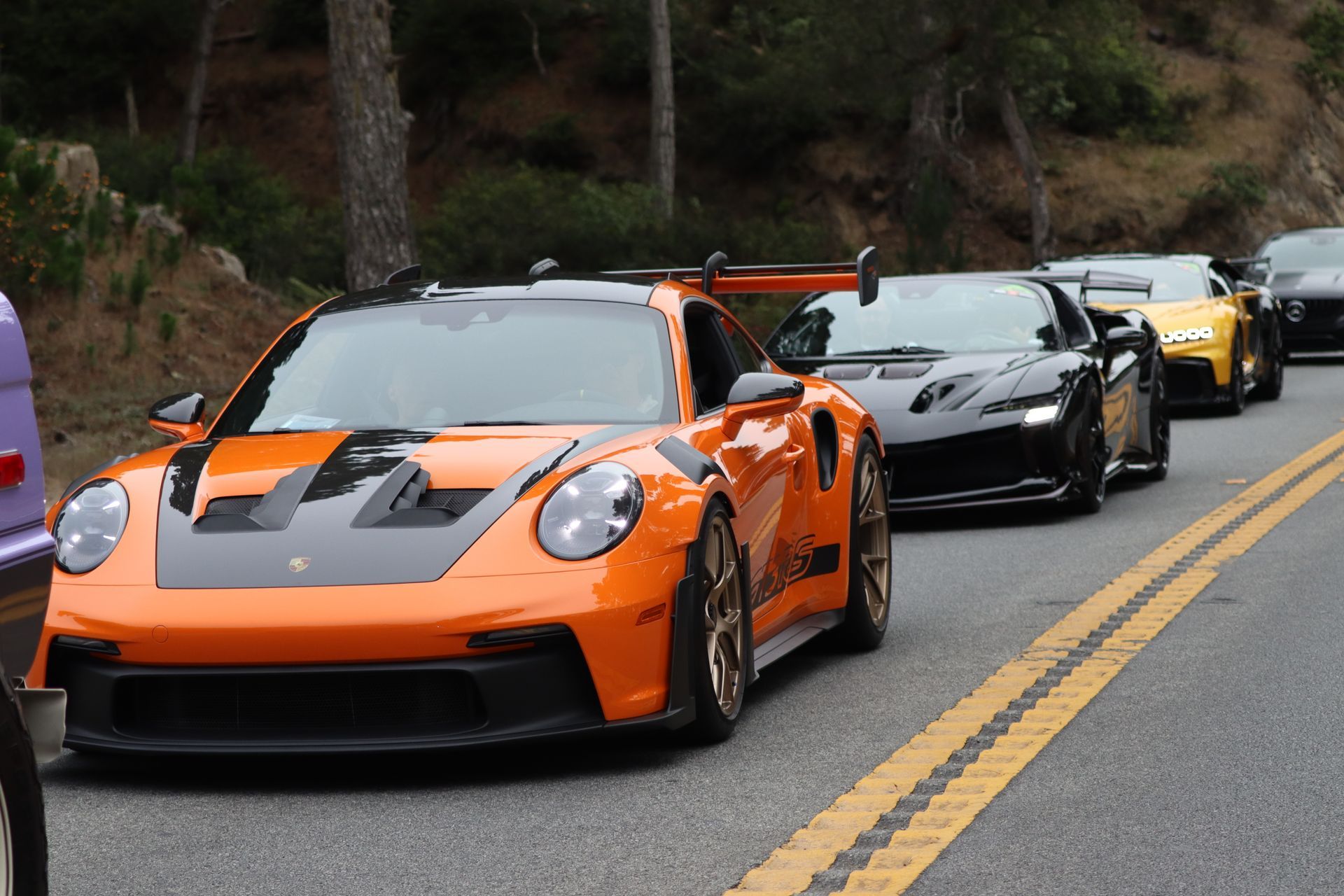 Line of sports cars on a mountain road, led by an orange Porsche with a black hood and spoiler. Line of sports cars on a mountain road, led by an orange Porsche with a black hood and spoiler.