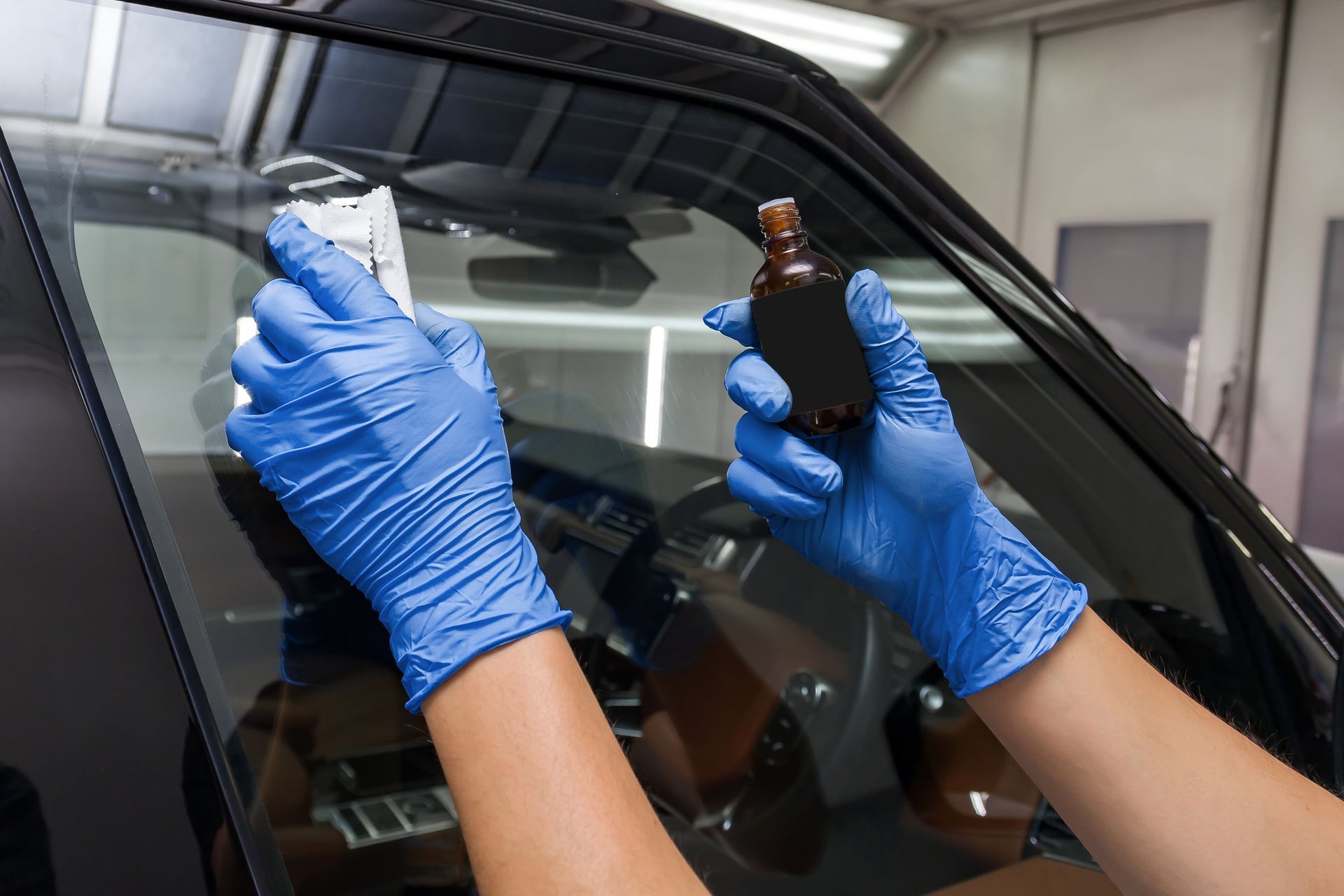 Blue-gloved hands cleaning a car windshield with a spray bottle in a workshop