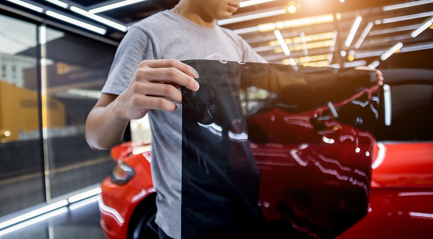 Person holding window tint film in front of a red car in a garage. Person holding up a black shirt in front of a red car in a garage