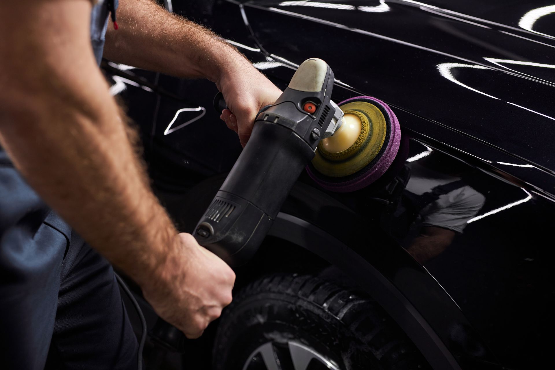 A person polishing a black car with a handheld buffer, creating a glossy shine.