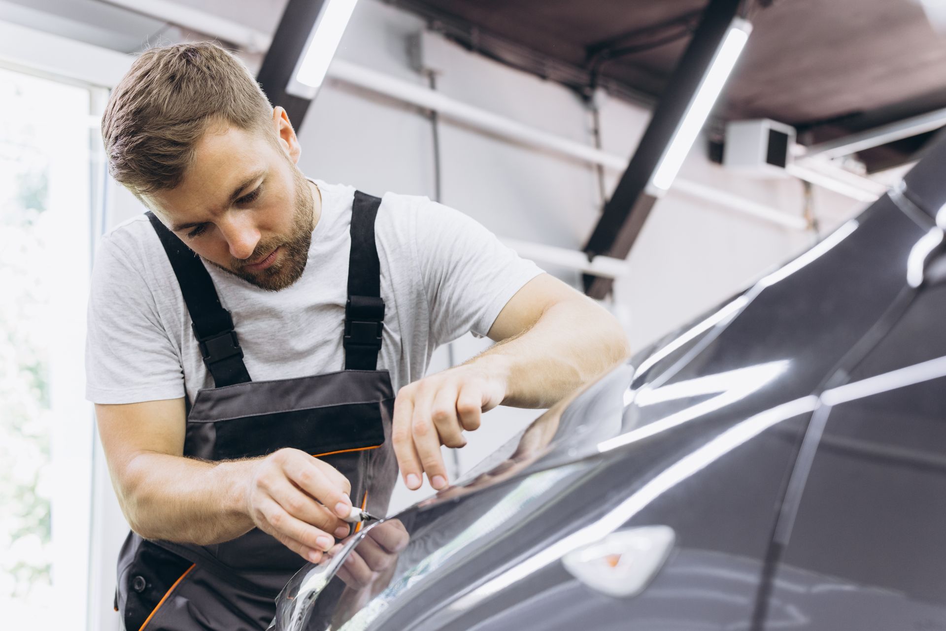 Male specialist cutting and wrapping a car in a garage showroom. Mechanic in gray shirt and overalls polishing a black car in a garage