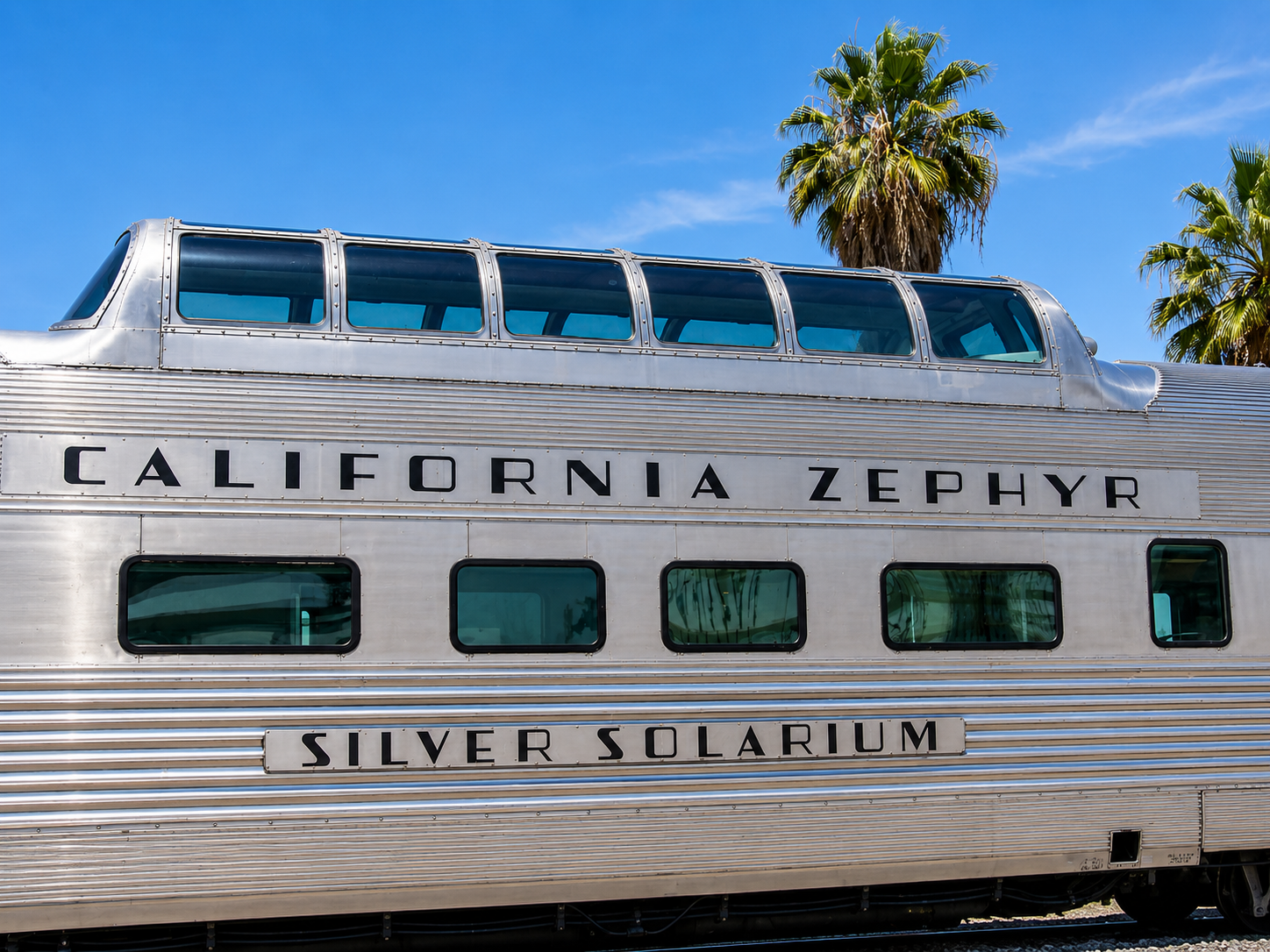 Silver California Zephyr train car with blue windows under a clear sky and palm trees
