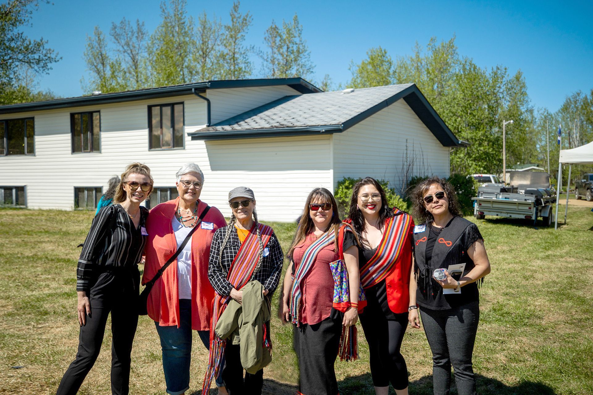 Six people smiling in front of a white building with a grassy yard on a sunny day.