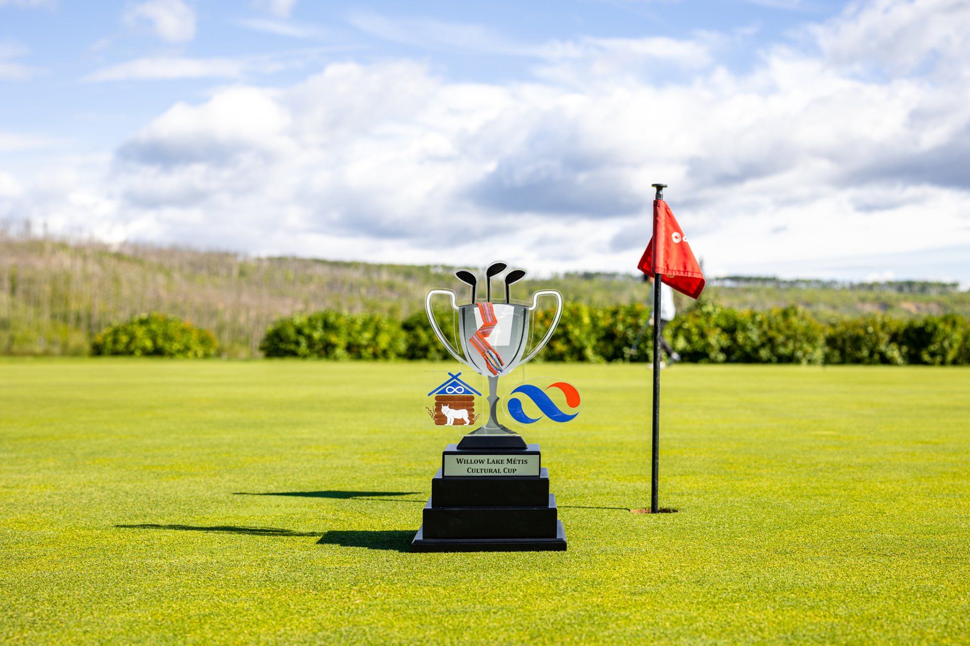 Golf trophy on a green field with a red flag, sunny sky in the background.