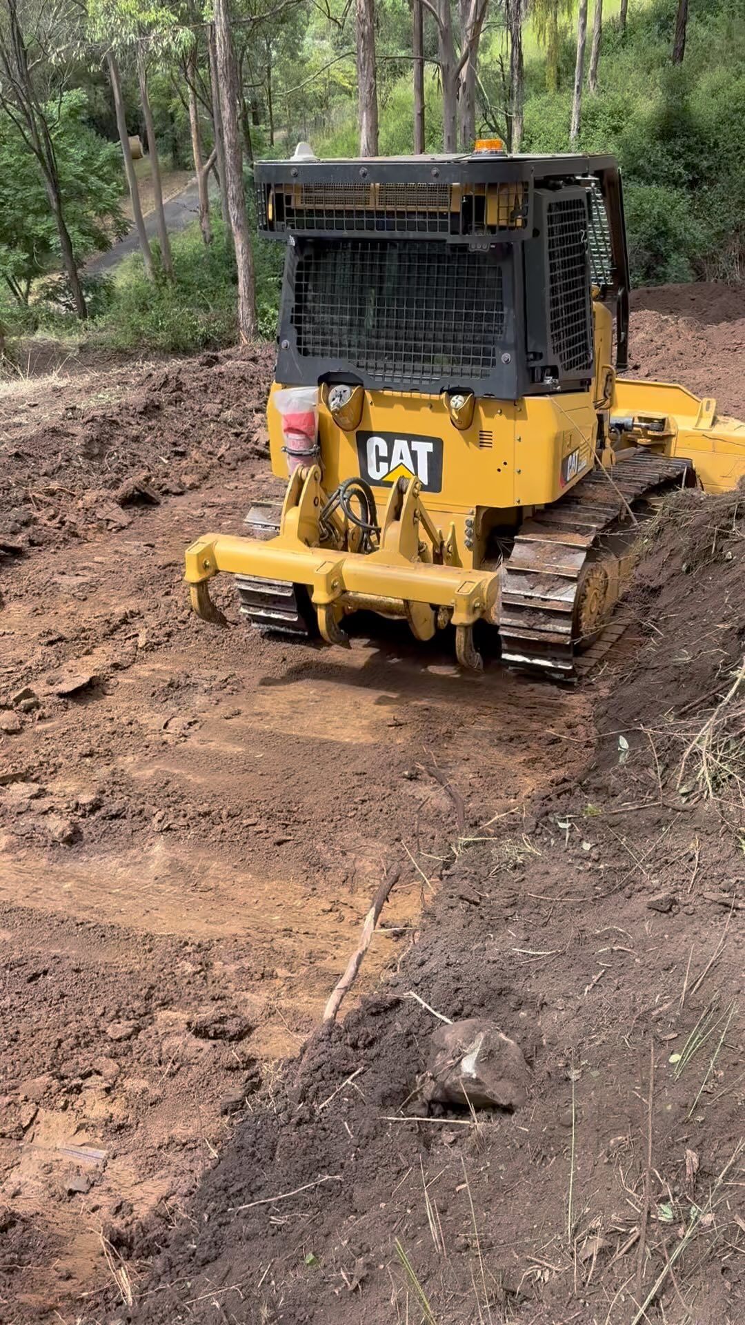 Yellow CAT bulldozer on a muddy dirt road in a wooded area, clearing and leveling the ground.