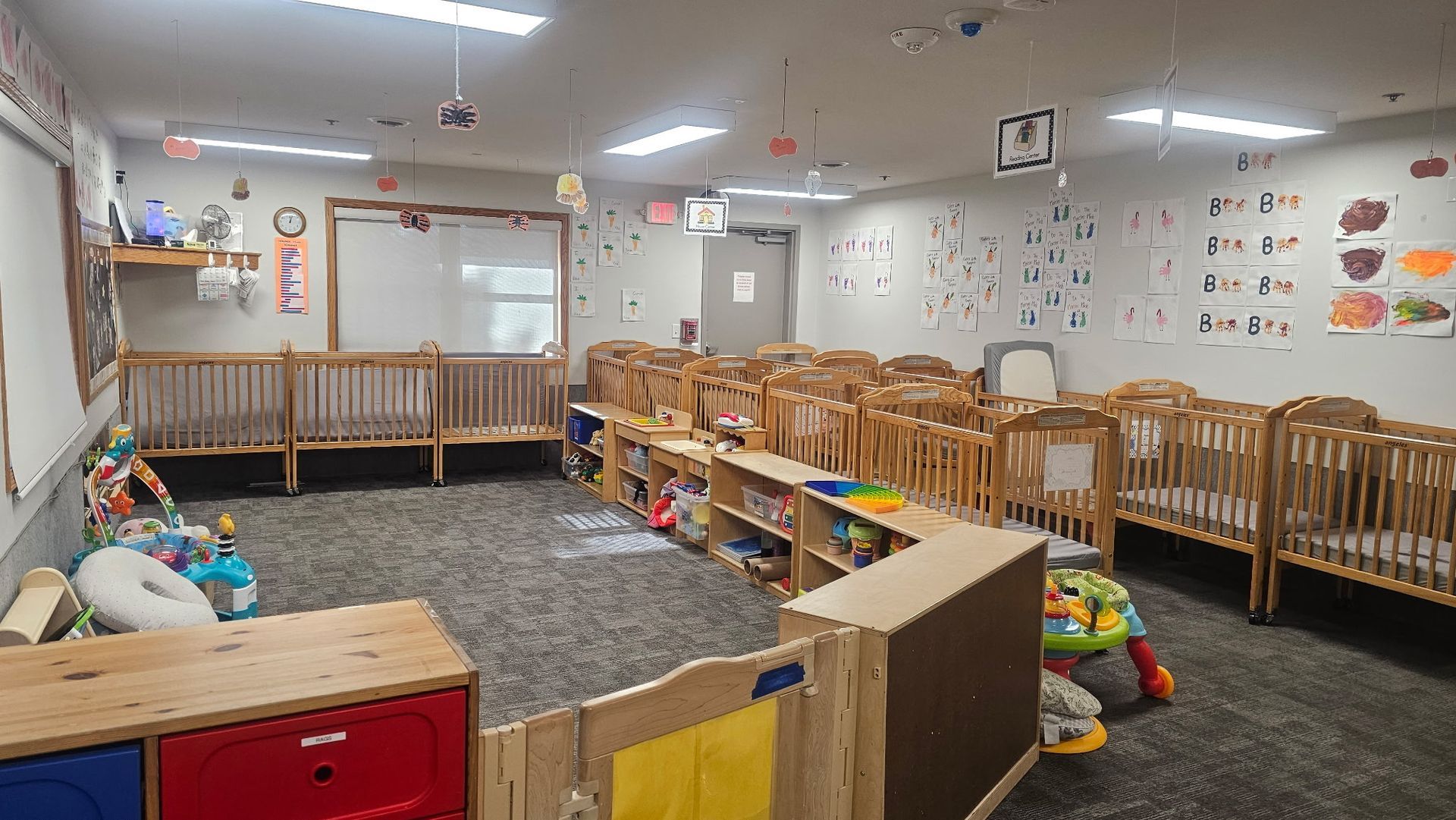 Daycare room with multiple cribs, shelving, toys, and educational decorations.