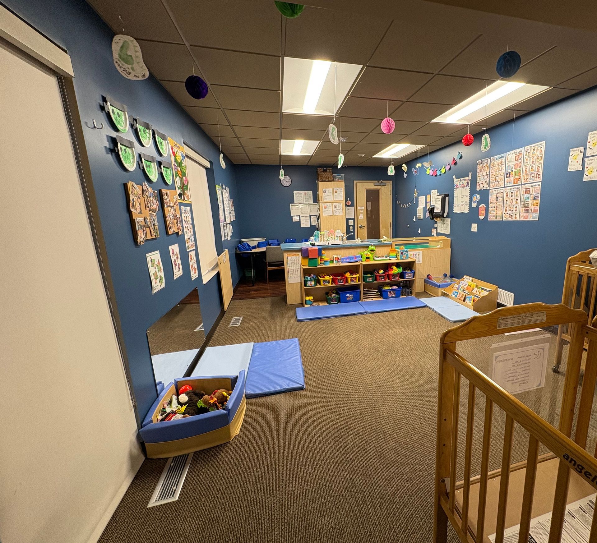 Childcare room, blue walls, toys, cribs, decorated with children's artwork and hanging decorations.