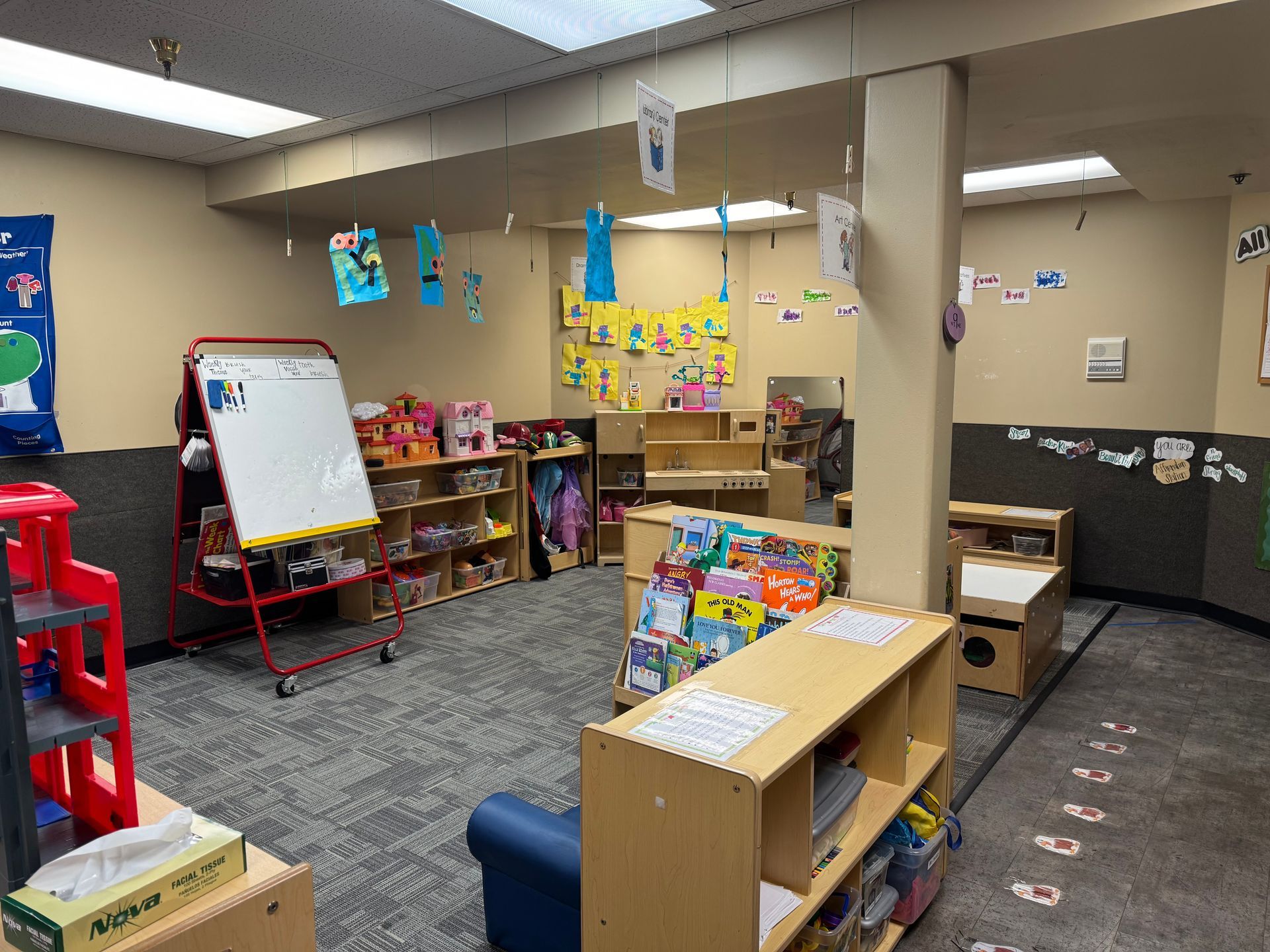 Children's classroom with shelves, toys, and art. A whiteboard and a blue sofa are visible.