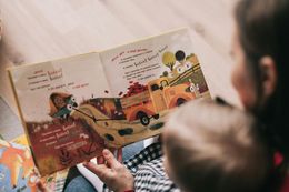 Adult reading a colorful picture book to a child; illustrations of a truck and landscape.