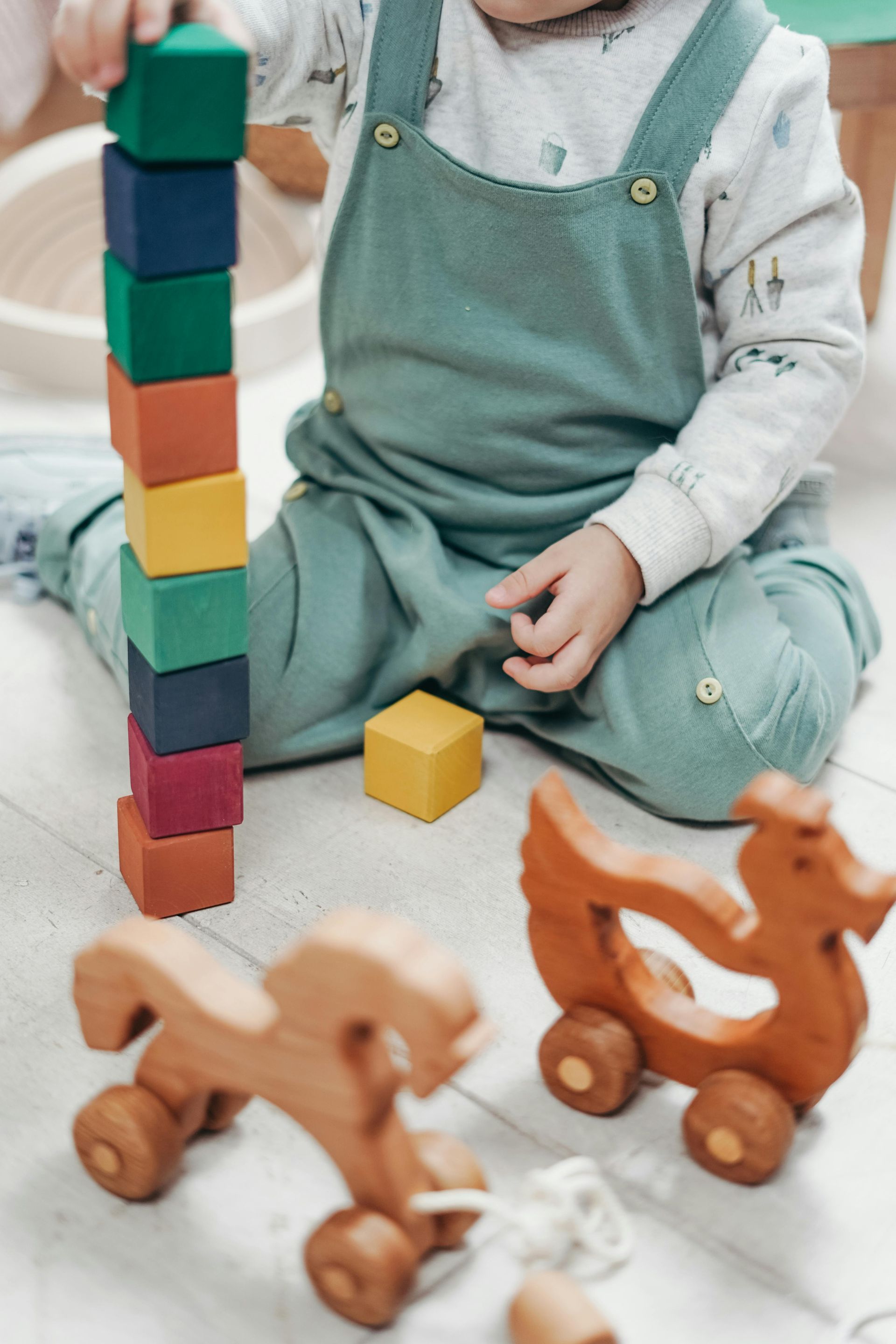 Child in green overalls stacking colorful blocks, wooden toys on floor.
