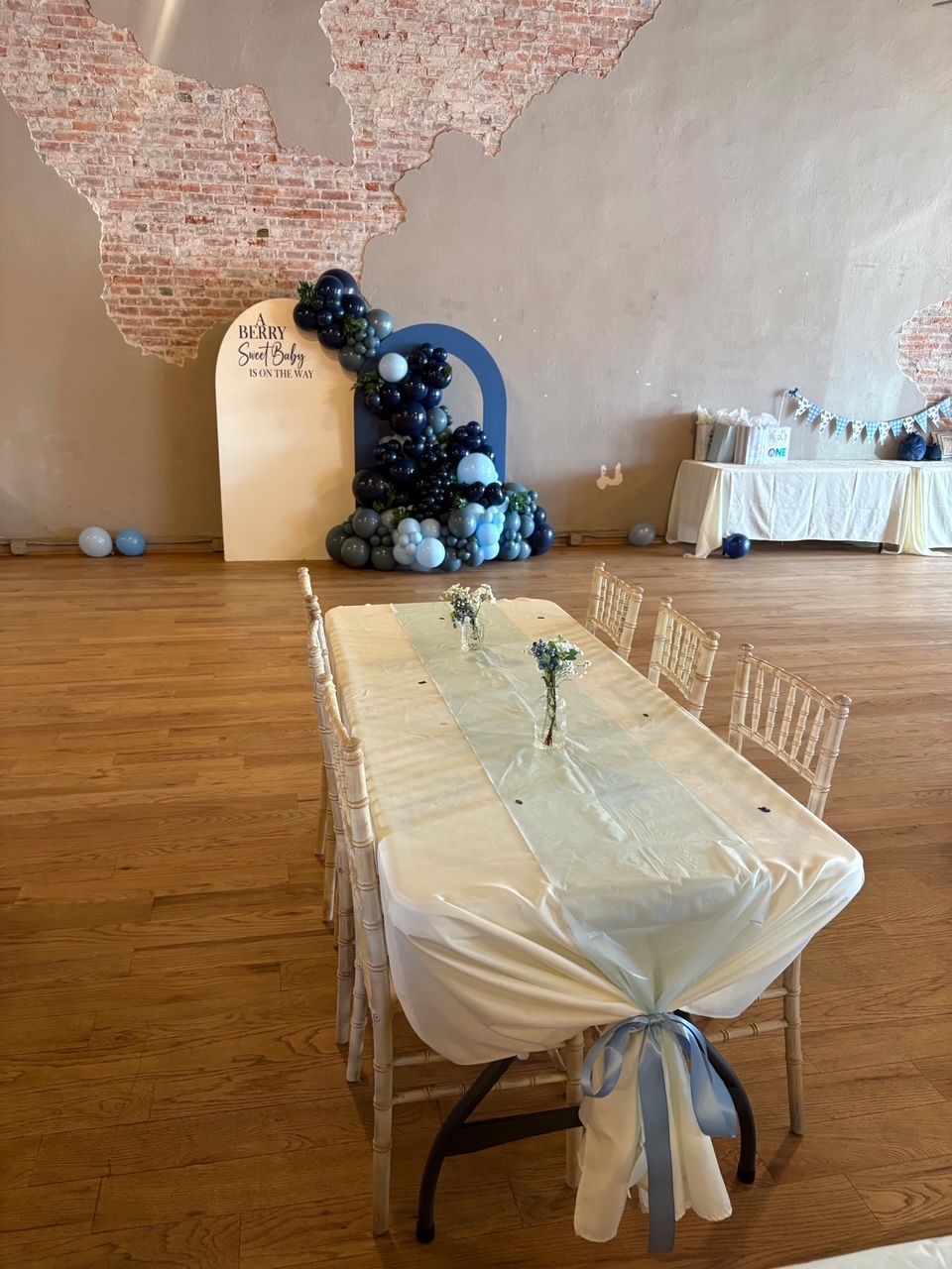 Decorated banquet table with white draping, blue balloon arch, and a small white cake in a rustic hall