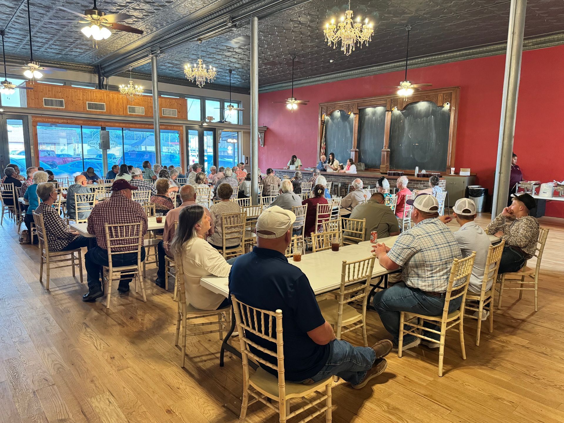 People seated at long tables in a bright restaurant or banquet hall with chandeliers and red walls.
