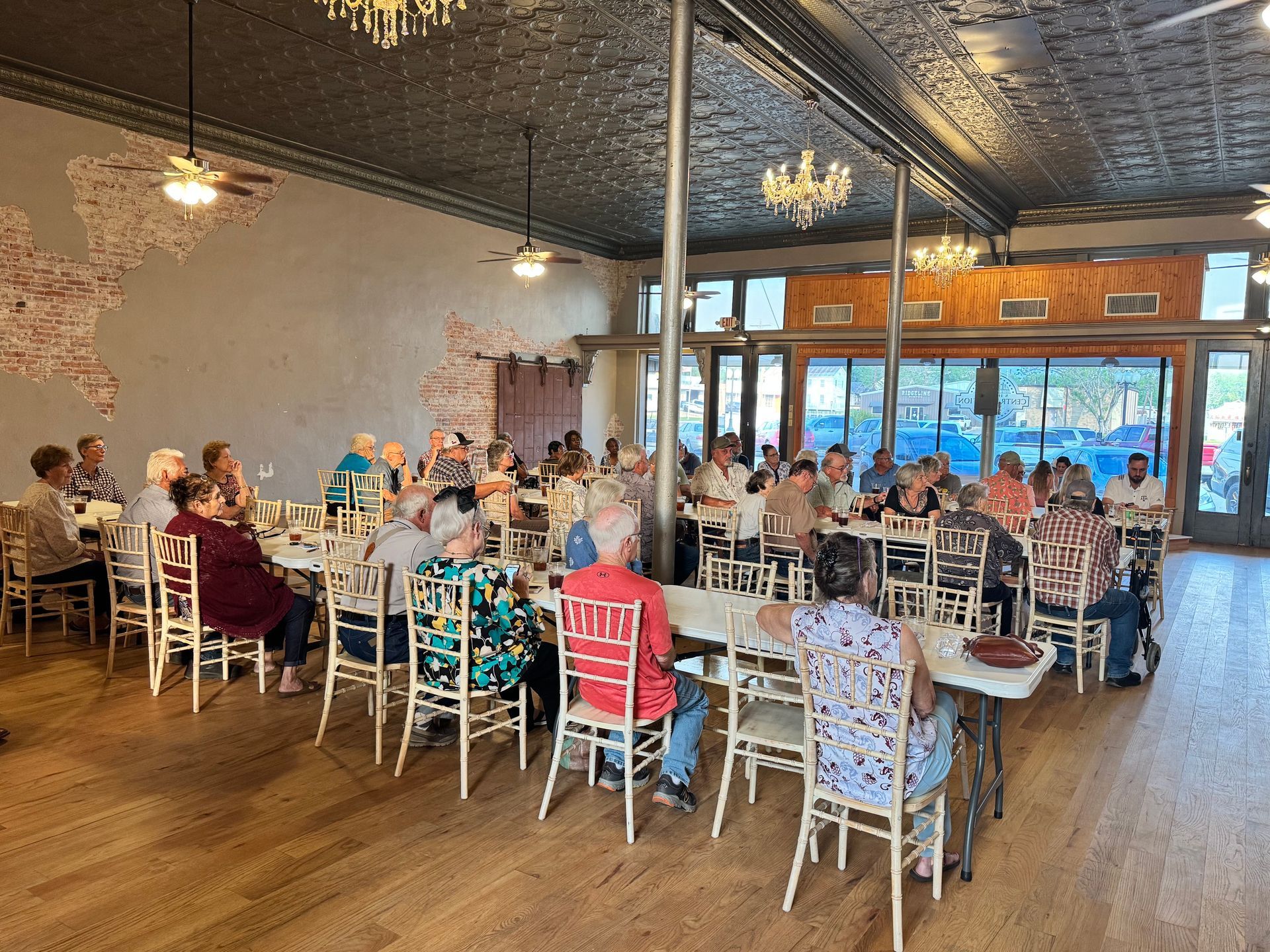 Guests seated at tables in a bright banquet hall with white chairs and hanging chandeliers