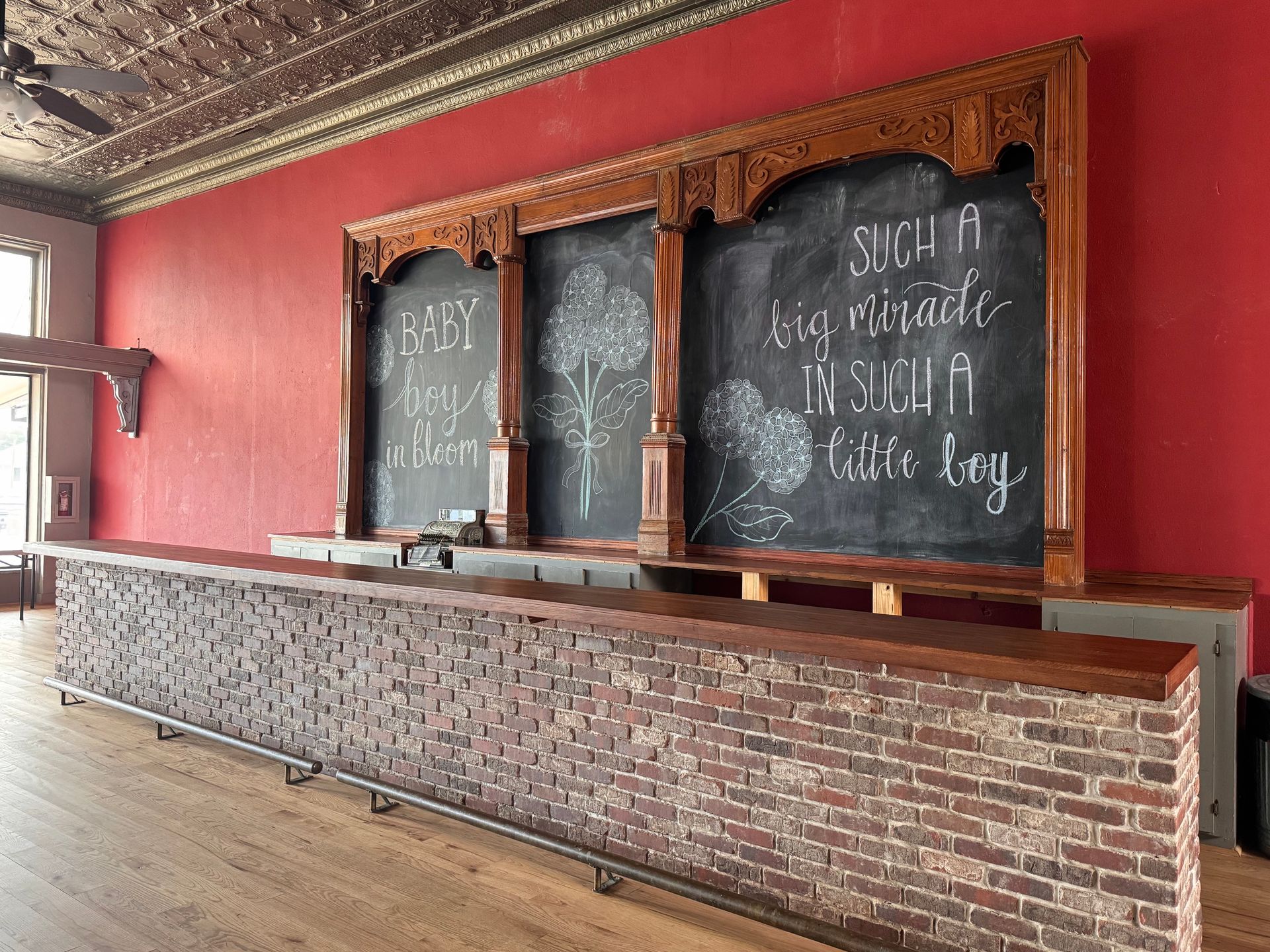 Empty bar interior with a long brick counter and three chalkboard menu panels on a red wall.