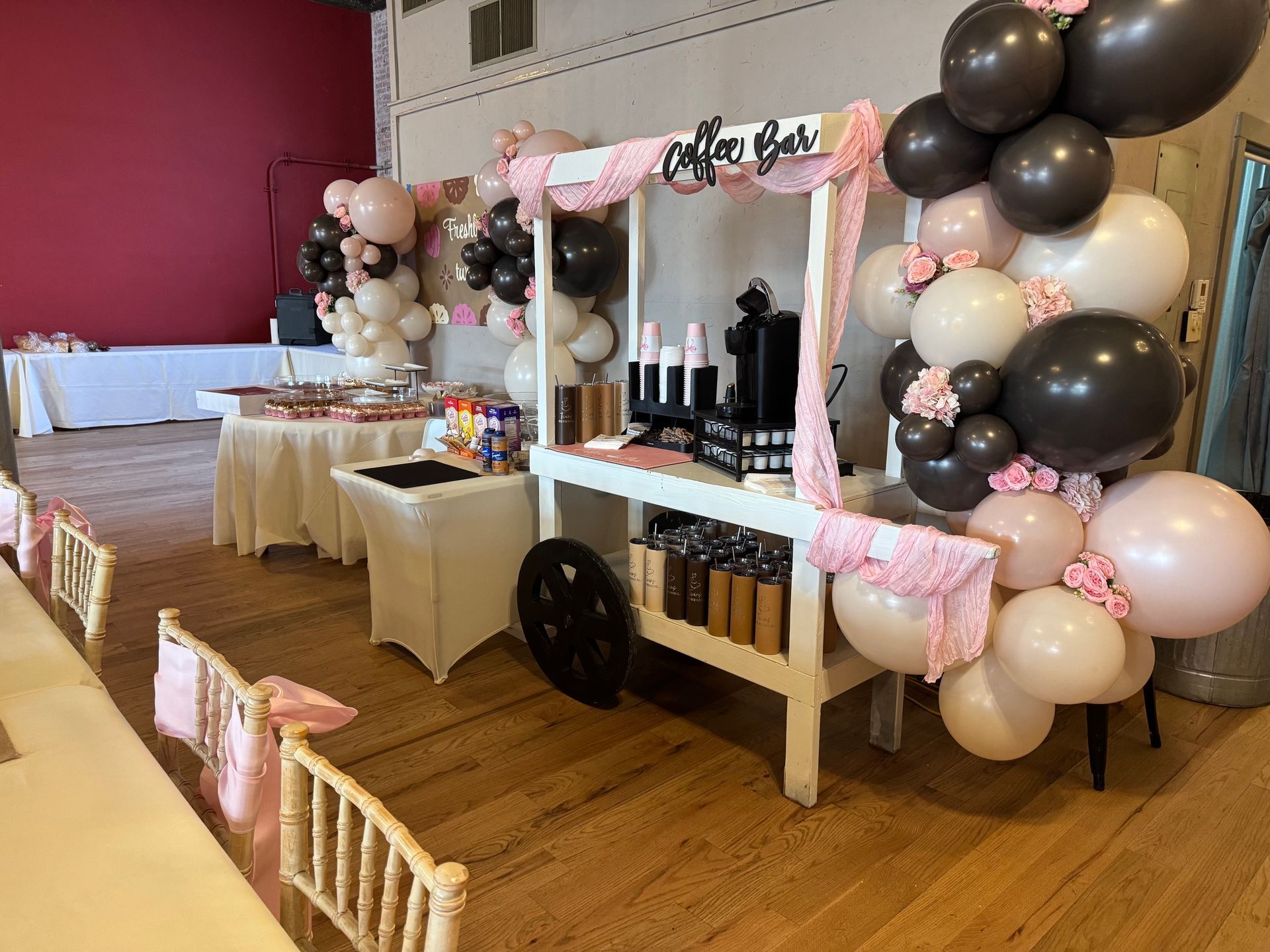 Pink-and-black dessert cart with balloons and bottles in a decorated event room