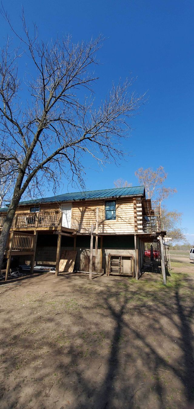 Rustic two-story house with a porch under a clear blue sky, framed by bare trees and a dirt yard.