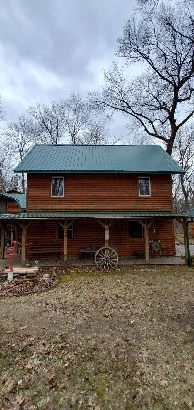 Rustic red cabin with green roof and porch in a leafless yard under a cloudy sky