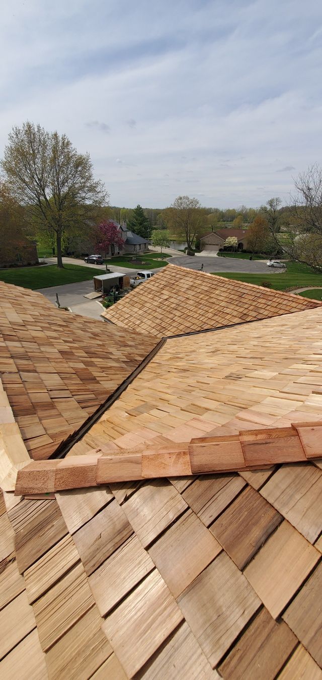 Rooftop with tan shingles and a central ridge, overlooking a green suburban landscape under a cloudy sky