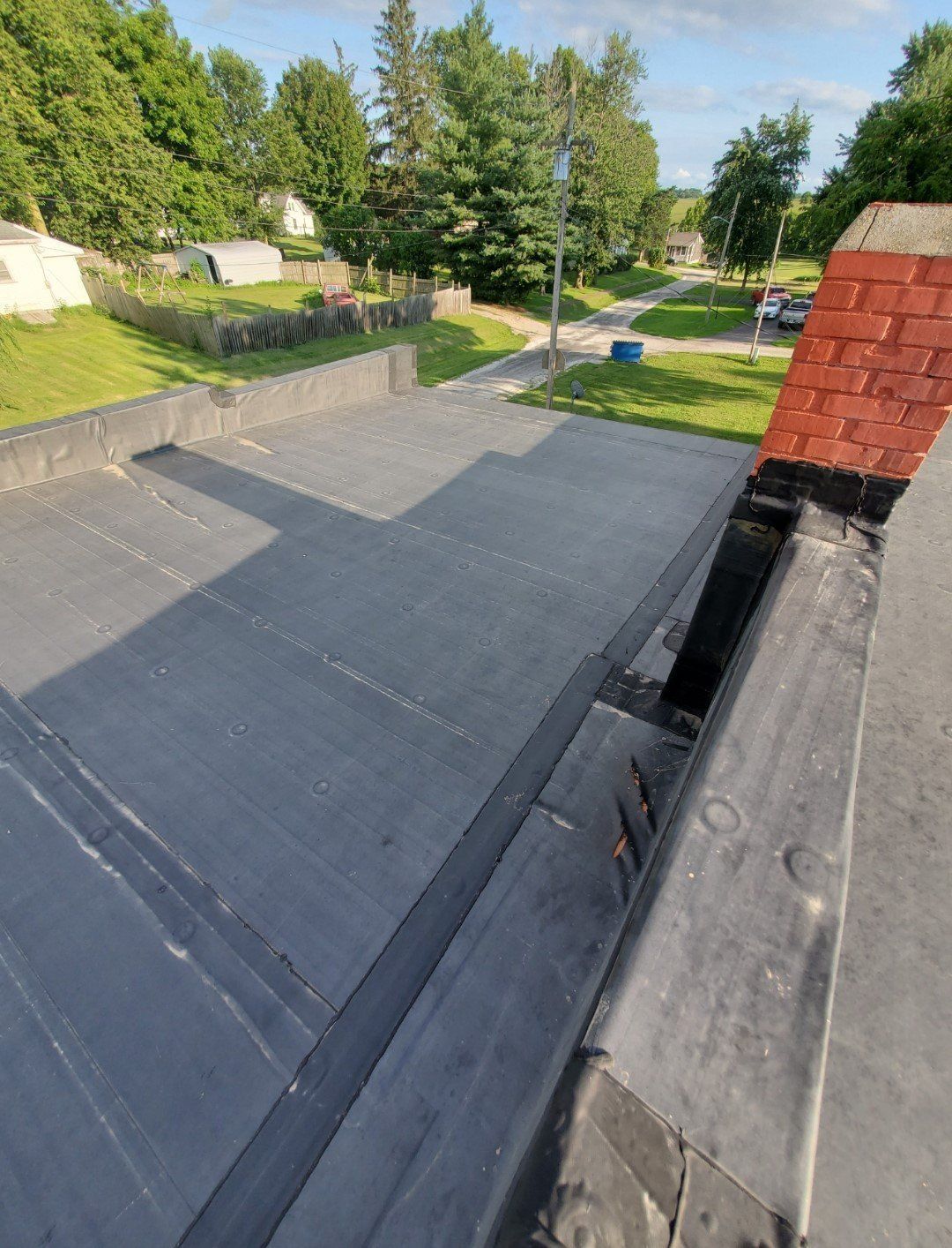 View from a rooftop over gray shingles toward a sunny neighborhood street and trees