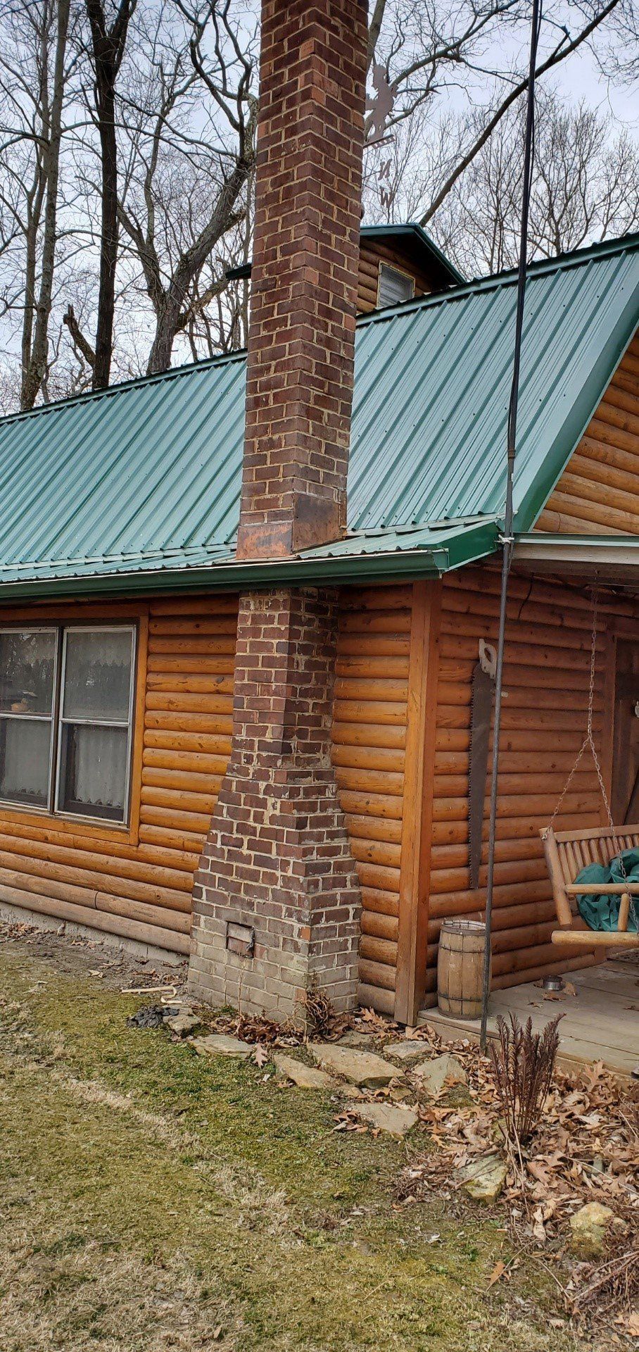 Log cabin with green metal roof and brick chimney in a wooded yard