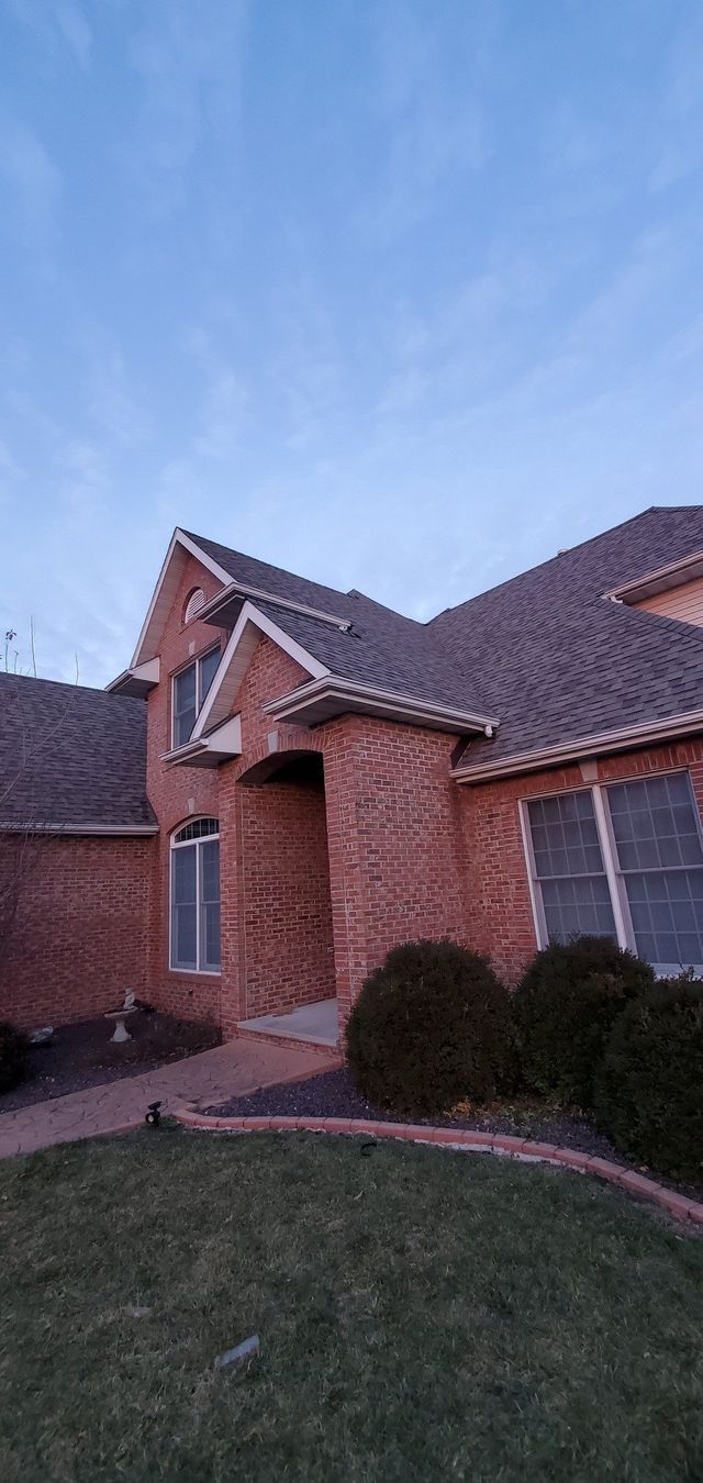 Brick suburban house with arched entry, dark roof, and trimmed bushes at dusk