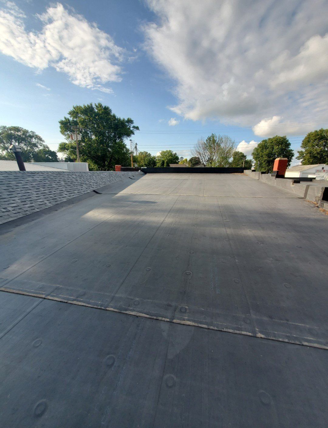 Empty flat rooftop with gray surface, low white wall, trees, and a partly cloudy blue sky.
