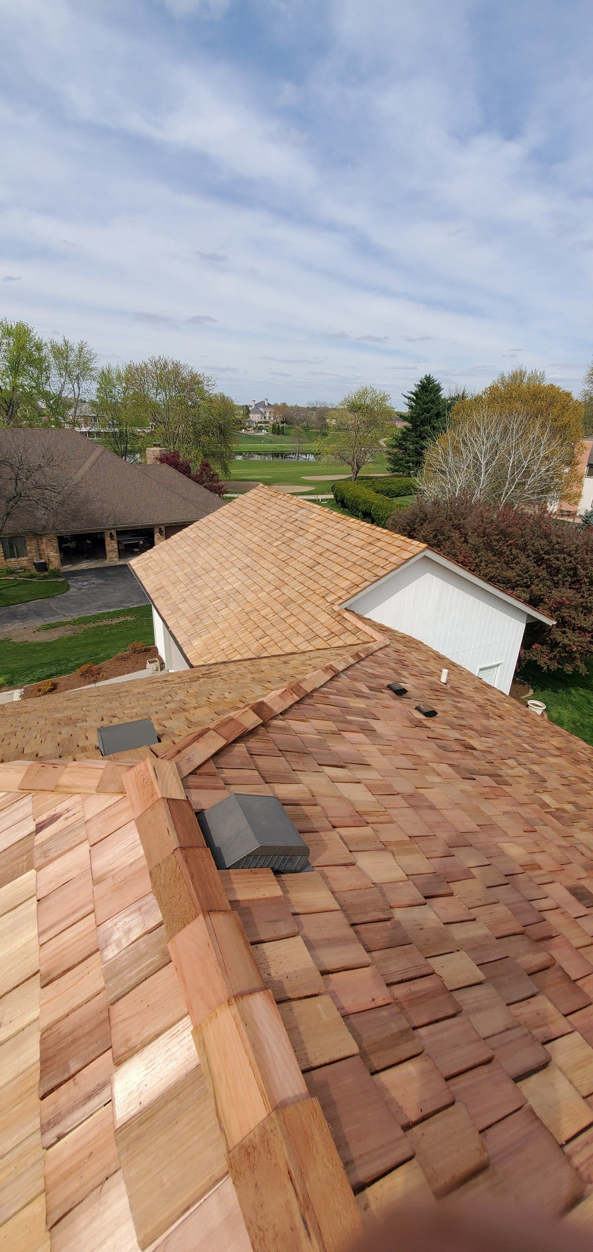 Rooftop view of tan shingles and a white dormer under a cloudy sky, with trees and a thatched roof nearby