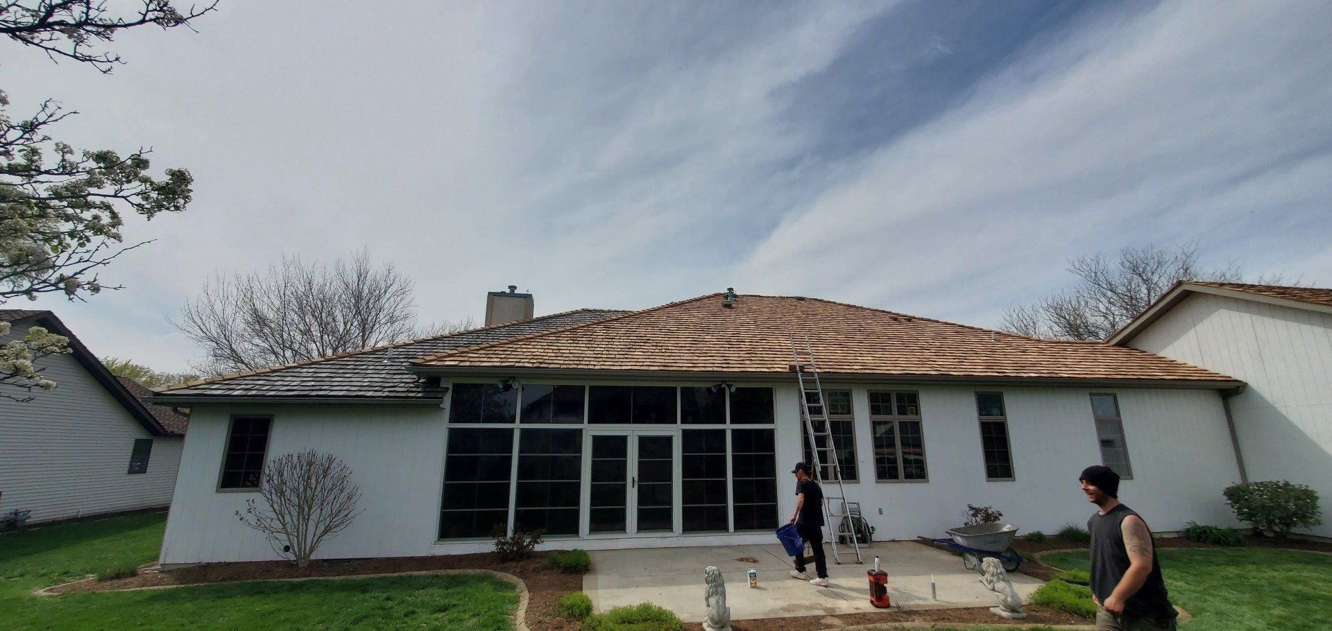 Single-story white house with damaged roof, people working outside under a cloudy sky