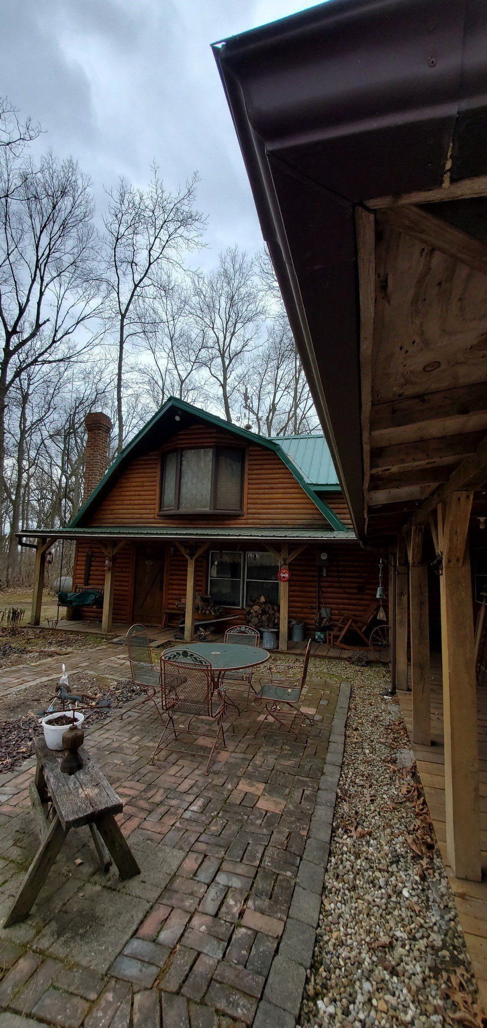 Rustic cabin with steep roof and wood siding in a wet brick courtyard, framed by bare trees and overcast sky