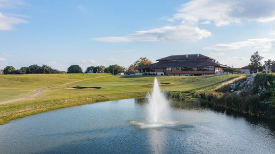 There is a fountain in the middle of a pond on a golf course.