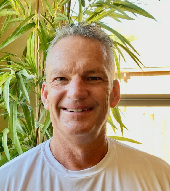 A man in a white shirt is smiling in front of a plant