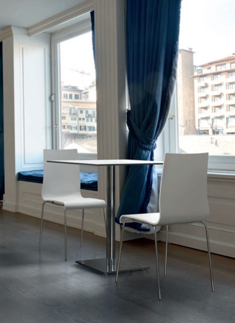 Due sedie bianche attorno a un tavolino accanto a finestre alte con tende blu e vista sulla città.