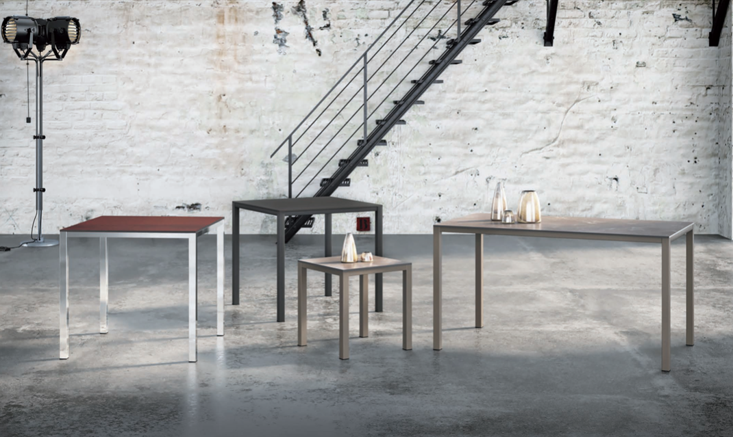 Loft in stile industriale con quattro semplici tavoli di diverse altezze, addossati a una parete di mattoni bianchi e a una scala.