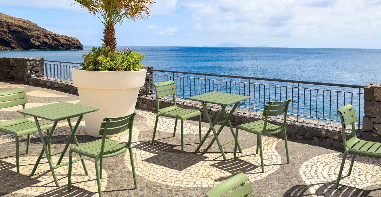 Terrazza esterna sul mare con tavoli e sedie verdi che si affacciano sull'oceano e sul cielo azzurro.