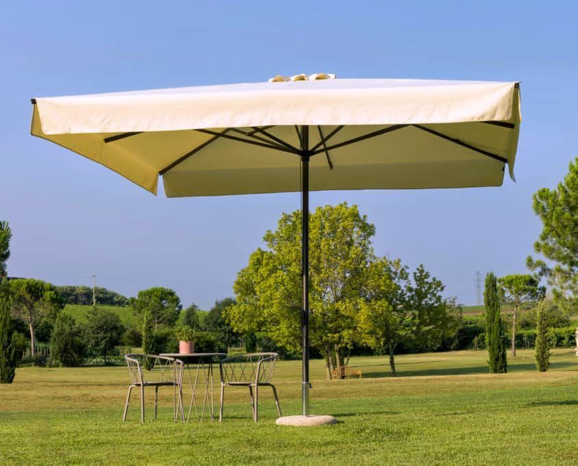Un grande ombrellone beige da giardino sovrasta un tavolo e sedie in metallo su un prato erboso con alberi sullo sfondo.
