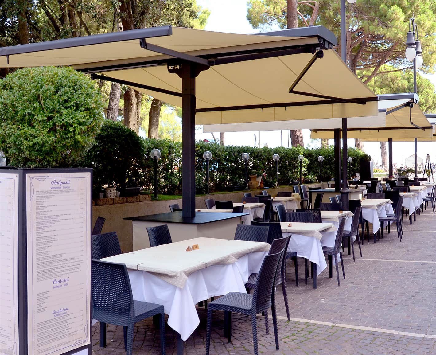 Terrazza esterna del caffè con tavolini con tovaglie bianche, ombrelloni beige e sedie vuote lungo un vialetto.