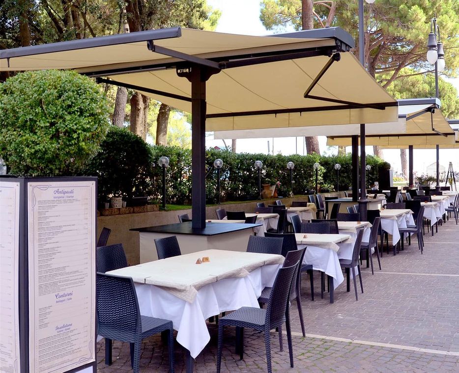 Terrazza esterna del ristorante con tavoli con tovaglie bianche, sedie nere e ombrelloni color cuoio lungo un vialetto.