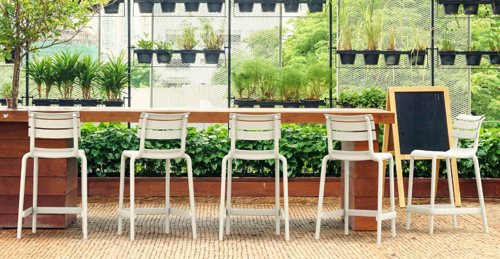 Terrazza esterna del caffè con sedie e tavoli bianchi, piante verdi e una lavagna con il menù vicino alla ringhiera.