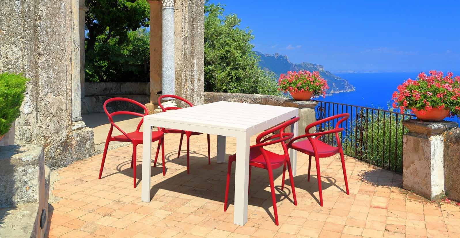 Tavolo da patio bianco con sedie rosse su una terrazza soleggiata con vista sul mare e sui fiori
