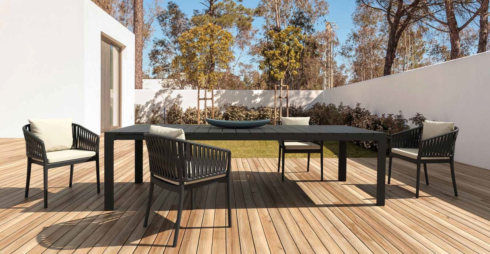 Patio moderno con mobili da esterno neri su una terrazza in legno, accanto a un muro bianco e ad alberi.