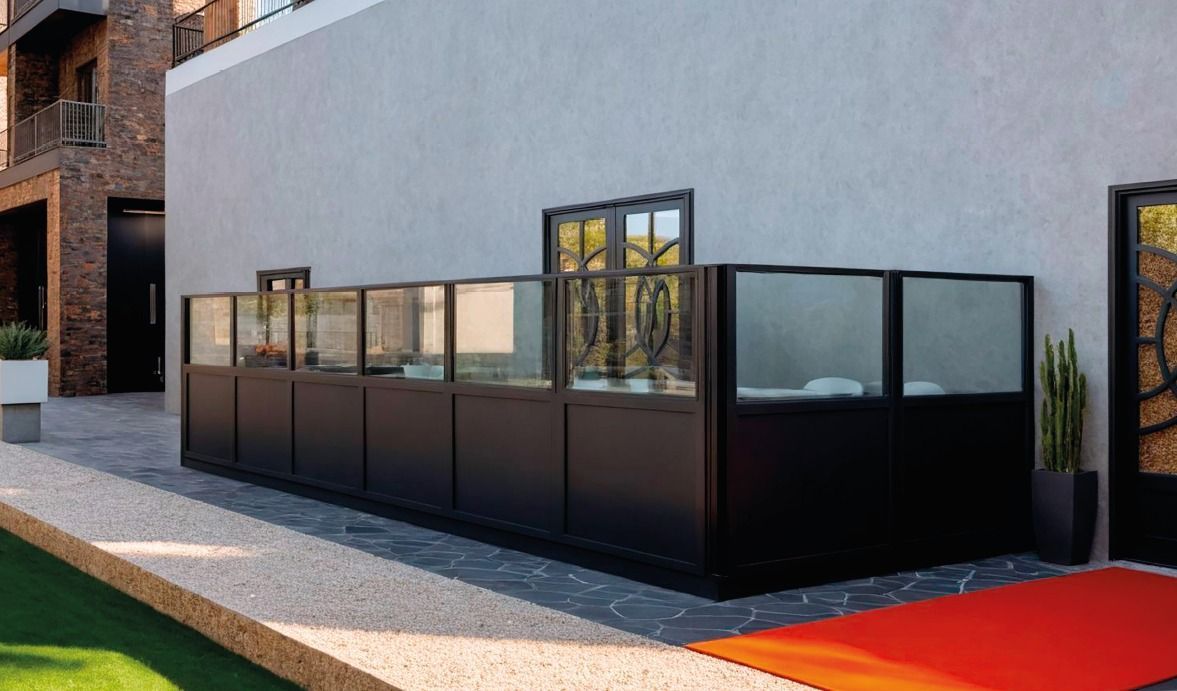 Patio esterno con pareti in vetro con telaio nero, adiacente a un edificio grigio e a un passaggio pedonale con pavimentazione rossa.